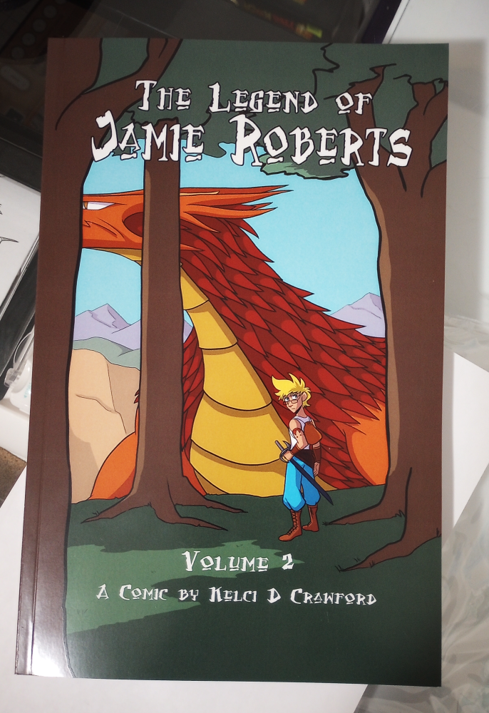 the printer's proof of The Legend of Jamie Roberts, volume 2, a comic by Kelci D Crawford. The cover depicts a blonde genderqueer person standing in a forest. Between the trees we see an enormous dragon with red and gold scales.