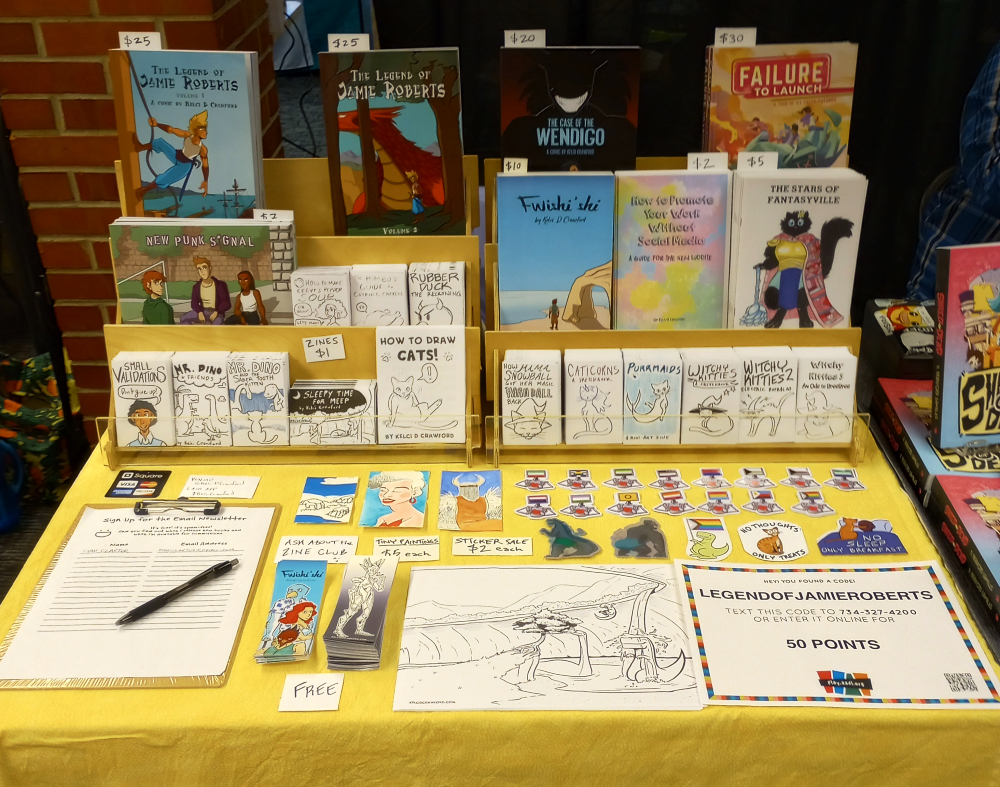 an assortment of comics, minicomics, zines, and stickers are on display on a bright yellow tablecloth. 